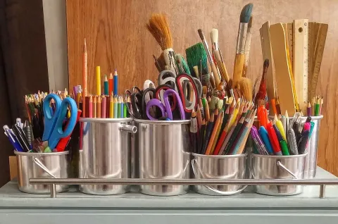 Brown background.  Silver containers holding art supplies such as paintbrushes, pencils, scissors, and markers.